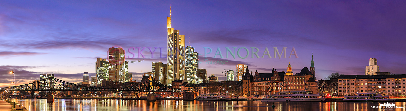 Panorama Bild Frankfurt am Main – Die abendlich beleuchtete Skyline von Frankfurt a. M. zusammen mit dem historischen Eisernen Steg im Vordergrund vom Mainufer in Frankfurt Sachsenhausen gesehen. Das Panorama ist kurz nach dem Sonnenuntergang, in der Dämmerung, bei einbrechender Nacht entstanden. 