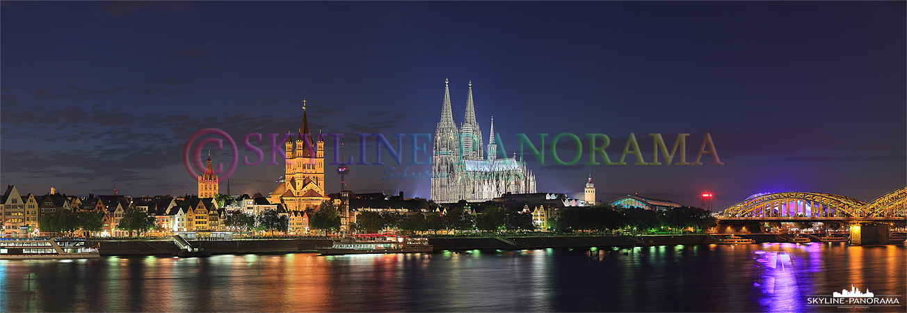 Die Kölner Skyline am Rheinufer vom Sockel der Deutzer Brücke aus gesehen. Das Köln Panorama zeigt den Rathausturm, die Kirche Sankt Martin und den Kölner Dom bei abendlicher Beleuchtung. 