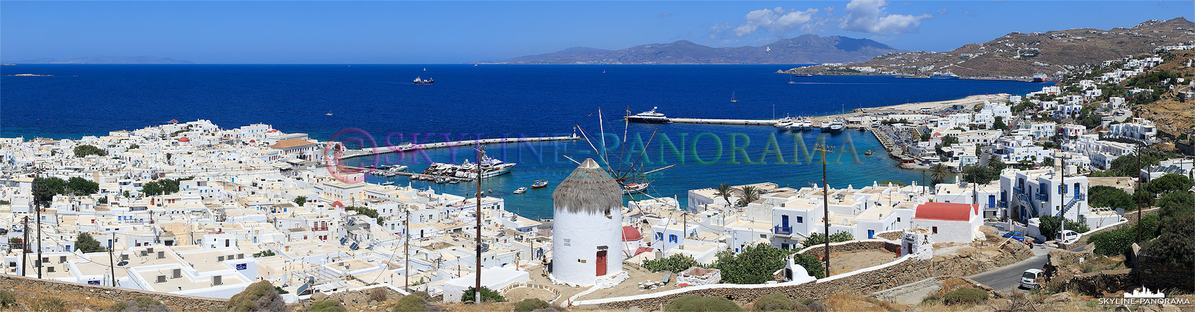 Panorama von dem oberhalb der Chora, auf dem Weg nach Ano Mera, gelegenen Aussichtspunkt auf die Windmühle Ano Myli und den Alten Hafen von Mykonos Stadt. 