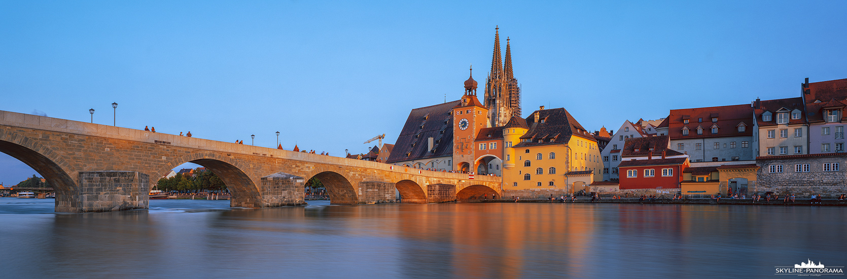 Die Altstadtansicht von Regensburg mit dem Regensburger Dom St. Peter, als bekanntestes Bauwerk der Stadt, und der Steinernen Brücke über der Donau. Das Panorama entstand zum Sonnenuntergang, wodurch die Fassaden in warmes Sonnenlicht getaucht sind.