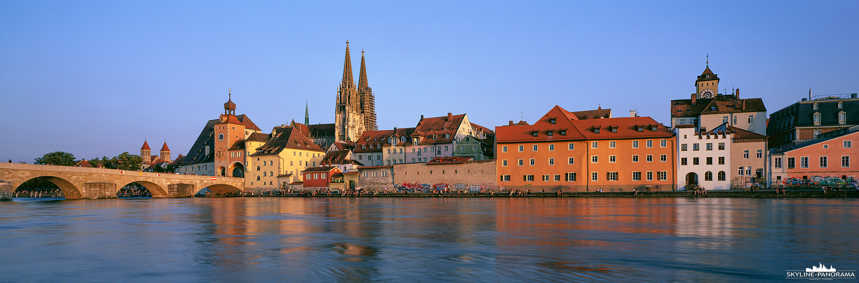 Die Silhouette der Altstadt von Regensburg als Panorama vom Ufer der Donau. Wie in vielen Städten der Welt, wird der Uferbereich in den Sommermonaten gern für einen abendlichen Sundowner genutzt, so auch hier in der bayrischen Metropole Regensburg.