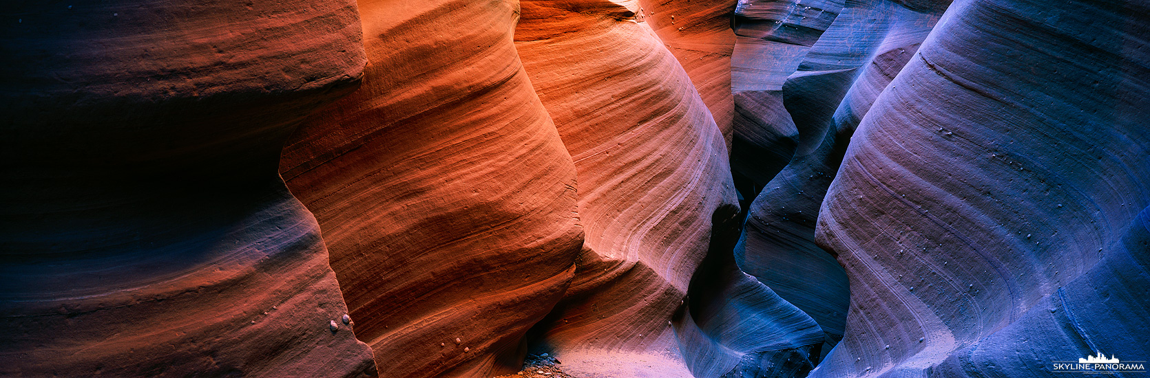 Ansicht einer Gesteinsformation im Slot Canyon des Watherhole Canyons in Page Arizona. Die unterschiedliche Färbung des Sandsteines ist dadurch zu erklären, dass der rechte Bereich des Panoramas durch einen kleinen Spalt von dem wolkenfreien Himmel beschienen wird und das verwendete Filmmaterial in diesem Fall nur das reflektierende Blau festhält. Film nimmt die Farben einer Lichtsituation auf einem anderen Weg war, als beispielsweise das menschliche Auge oder der Sensor eine Digitalkamera. Der linke Bereich des Panoramas wird hingegen direkt von der Nachmittagssonne beschienen, wodurch der Sandstein im warmen Licht strahlt.